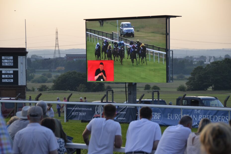 A crowd of people watching a horse race on a big screen with a BSL interpreter